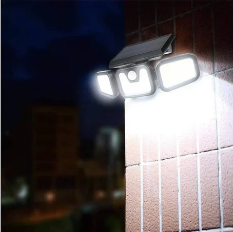 Na zdjęciu widoczna jest zewnętrzna lampa LED z panelem solarnym, zamontowana na ścianie budynku. Lampa składa się z trzech ruchomych paneli świecących intensywnym, białym światłem, oświetlając okolicę po zmroku. W tle widać rozmazane kontury innych budynków nocą.
