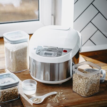 Biało-srebrny multicooker ADLER ustawiony na blacie kuchennym, otoczony pojemnikami z ryżem, płatkami owsianymi i orzechami